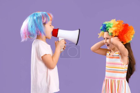 Foto de Hermano pequeño y hermana con pelucas de colores y megáfono sobre fondo lila. Celebración del Día de los Inocentes - Imagen libre de derechos