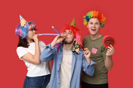 Junge Freunde in lustiger Verkleidung mit Trillerpfeifen und Pfeifenkissen auf rotem Hintergrund. Aprilscherz-Feier