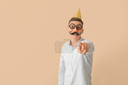 Foto de Hombre de negocios disfrazado de gracioso apuntando al espectador sobre fondo beige. Celebración del Día de los Inocentes - Imagen libre de derechos