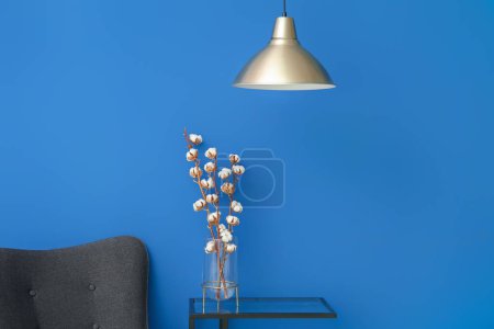Vase of cotton sprigs on glass table with lamp and armchair near blue wall