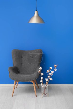 Vase of cotton sprigs with lamp and armchair near blue wall