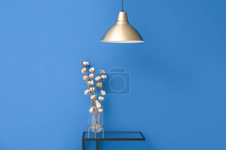Vase of cotton sprigs on glass table with lamp near blue wall