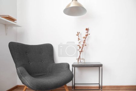 Vase of cotton sprigs on black table with lamp and armchair near white wall