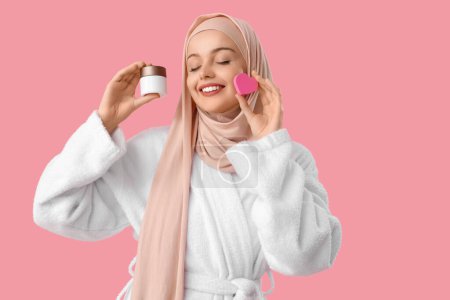 Beautiful Muslim woman in hijab with heart-shaped sponge and cream on pink background