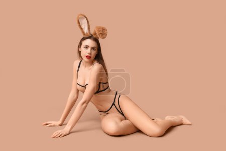 Sexy young woman in bunny ears sitting on beige background. Easter celebration