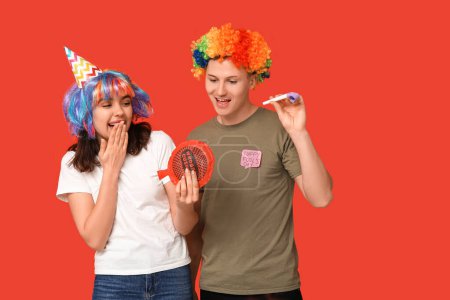 Young friends in funny disguise with party whistle and whoopee cushion on red background. April fool's day celebration