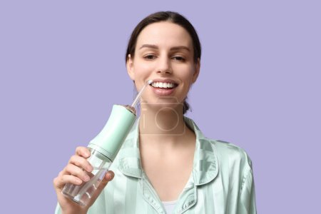 Schöne junge Frau mit oralem Bewässerungsgerät auf fliederfarbenem Hintergrund