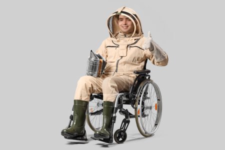 Apicultor masculino en silla de ruedas con ahumador mostrando pulgar hacia arriba sobre fondo claro