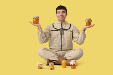 Male beekeeper with jars of honey sitting on yellow background