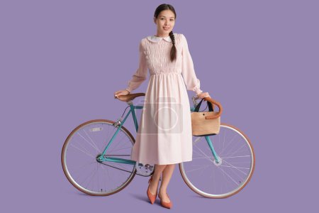 Foto de Hermosa mujer asiática joven con bicicleta y bolso elegante sobre fondo lila - Imagen libre de derechos