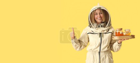 Female beekeeper with jars of sweet honey showing thumb-up gesture on yellow background with space for text