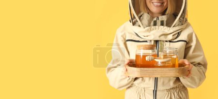 Female beekeeper with jars of sweet honey on yellow background with space for text