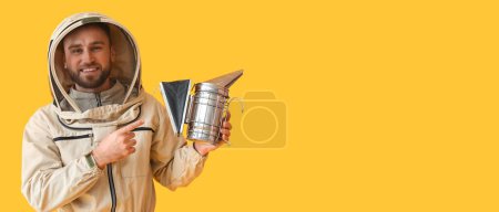 Male beekeeper in protective suit pointing at smoker on yellow background with space for text
