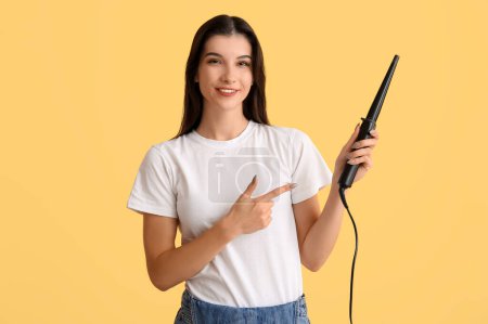 Photo for Beautiful young happy woman pointing at curling iron on yellow background - Royalty Free Image