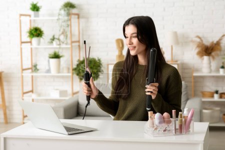 Photo for Beautiful young happy female blogger with laptop and curling iron recording video at home - Royalty Free Image