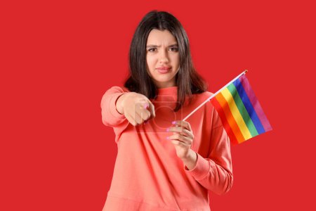 Mujer joven con bandera LGBT señalando al espectador sobre fondo rojo. Concepto de acusación