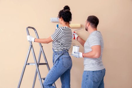 Foto de Hermosa pareja joven con rodillos de pintura cerca de la pared beige haciendo reparación en casa nueva - Imagen libre de derechos