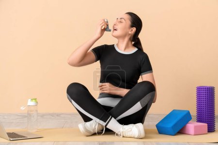 Young woman with sports equipment using inhaler near beige wall