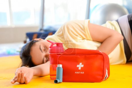 Inhalador con botiquín de primeros auxilios, botella de agua y mujer inconsciente en gimnasio, primer plano