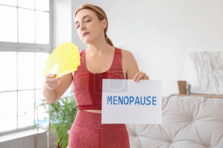 Deportiva mujer madura sosteniendo papel con palabra MENOPAUSE y abanico en casa