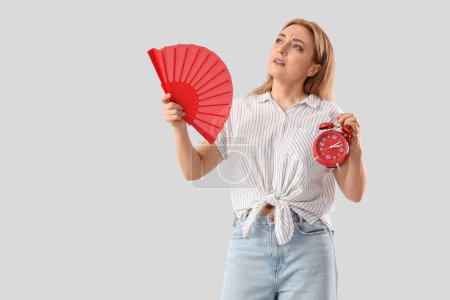 Mature woman with alarm clock and fan on light background. Menopause concept