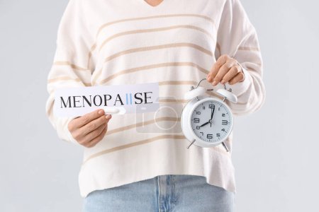 Mature woman holding paper with word MENOPAUSE and alarm clock on light background, closeup