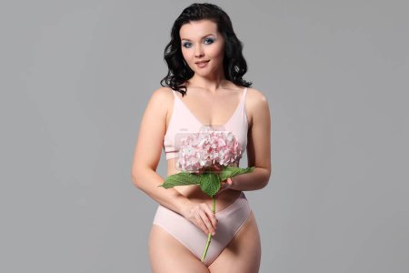 Beautiful young body positive woman in stylish underwear with hydrangea flowers on grey background