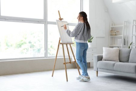 Young woman with dreadlocks painting on easel at home