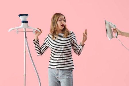 Hübsche junge Frau bevorzugt modernen Kleidungsdampfer vor elektrischem Bügeleisen auf rosa Hintergrund
