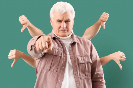 Photo for Mature man pointing at viewer and people showing thumbs-down on green background. Accusation concept - Royalty Free Image