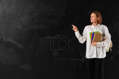 Photo for Female student with book and copybooks pointing at something on blackboard background. End of school concept - Royalty Free Image