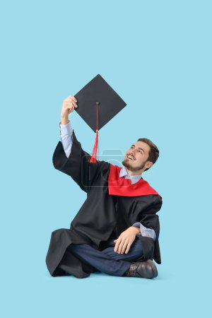 Photo for Male graduate student with mortar board sitting on blue background - Royalty Free Image