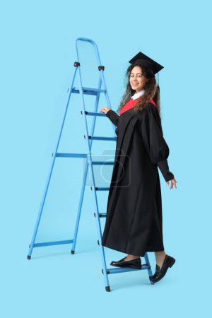 Photo for African-American female graduate student climbing stepladder on blue background - Royalty Free Image