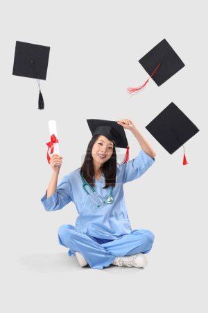Asian female medical graduate student with diploma and falling mortar boards on white background