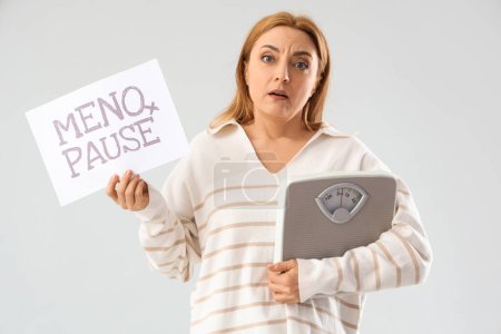 upset mature woman holding paper with word MENOPAUSE and weight scales on light background