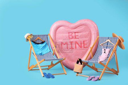 Inflatable mattress in shape of heart with text BE MINE, sun lounger and swimsuits on blue background