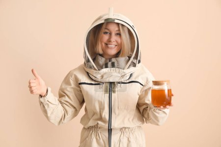Female beekeeper with jar of sweet honey showing thumb-up gesture on beige background