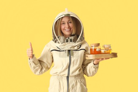 Female beekeeper with jars of sweet honey showing thumb-up gesture on yellow background