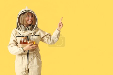 Female beekeeper with jars of sweet honey pointing at something on yellow background