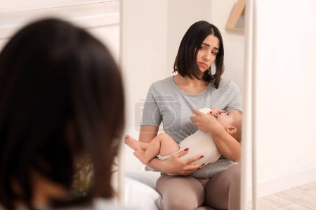 Junge Frau mit Baby, das an postnataler Depression leidet, in der Nähe des Spiegels im Schlafzimmer