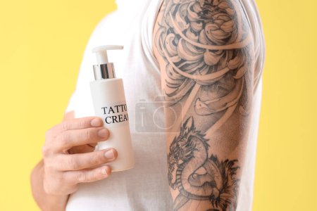 Young man with bottle of tattoo cream on yellow background, closeup