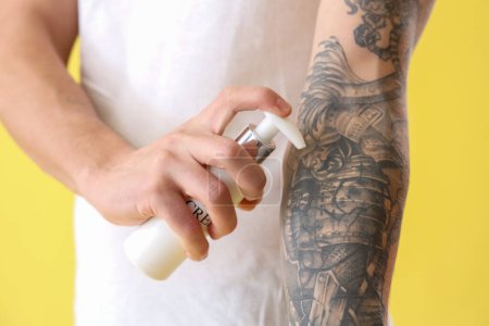 Hombre joven aplicando crema de tatuaje sobre fondo amarillo, primer plano