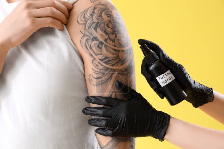 Tattoo master spraying man's arm against yellow background, closeup