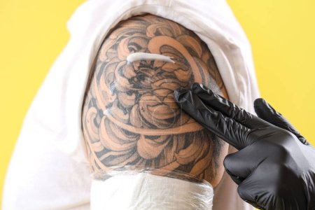Tattoo master applying cream on man's shoulder against yellow background, closeup