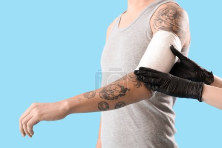 Maestro aplicando película protectora del tatuaje en el brazo del hombre sobre fondo azul, primer plano