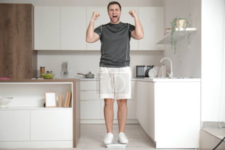 Téléchargez les photos : Heureux jeune homme sportif mesurant le poids sur des balances dans la cuisine - en image libre de droit