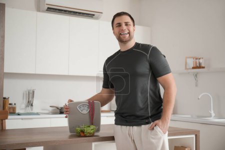 Téléchargez les photos : Jeune homme sportif avec balances, ruban à mesurer et salade sur la table dans la cuisine. Concept de perte de poids - en image libre de droit