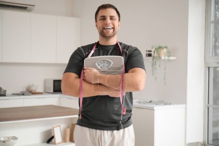 Téléchargez les photos : Jeune homme sportif avec ruban à mesurer et balances dans la cuisine. Concept de perte de poids - en image libre de droit