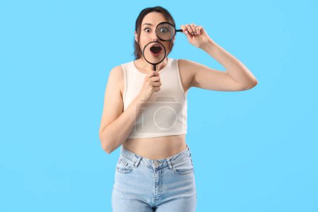 Schockierte junge Frau mit Lupe auf blauem Hintergrund