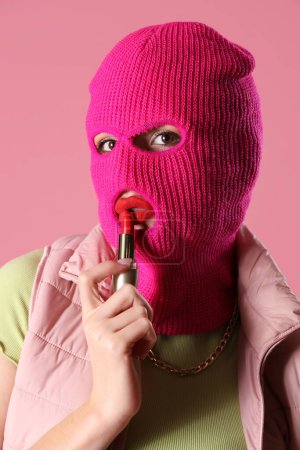 Schöne junge stilvolle Frau mit Sturmhaube, die roten Lippenstift auf rosa Hintergrund aufträgt